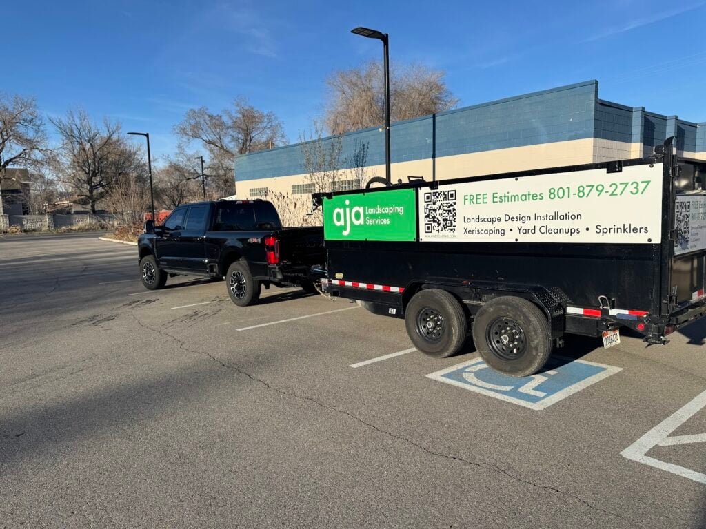 A black pickup truck towing an AJA Landscaping services trailer is parked in an empty lot near a blue building on a clear day, showcasing the company’s commitment to quality that’s reflected in positive customer feedback.