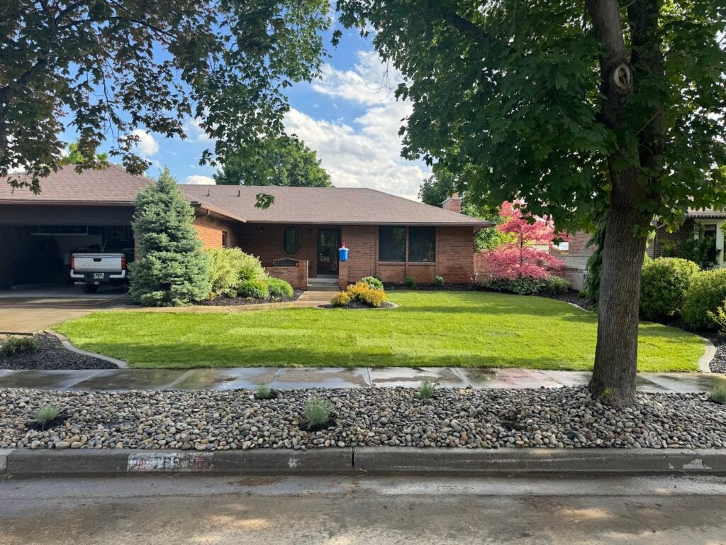Single-story brick house with an attached garage, well-kept lawn, trees, shrubs, and a rock-bordered flowerbed along the sidewalk under a partly cloudy sky—showcasing thoughtful landscape design in this charming West Jordan, Utah neighborhood.