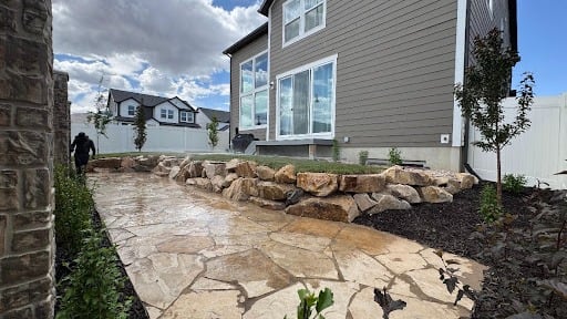 A stone walkway runs alongside a modern house with large windows, bordered by a low rock retaining wall, young plants, and a grassy area under a cloudy sky—showcasing the quality and attention to detail that have earned AJA Landscaping positive customer reviews.