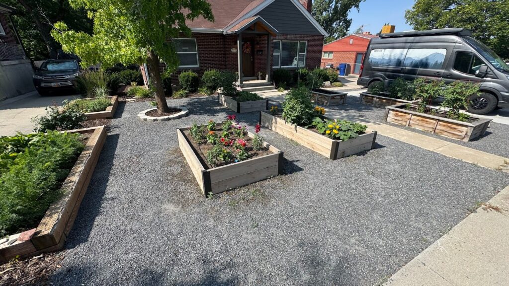 Front yard with multiple raised garden beds containing various plants and flowers, surrounded by gravel paths, in front of a brick house and a parked van.