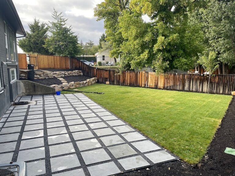 Backyard with a tiled patio, green lawn, wooden fence, stone retaining wall, and large trees in the background—an inviting space enhanced by expert hardscape design for lasting beauty and function.