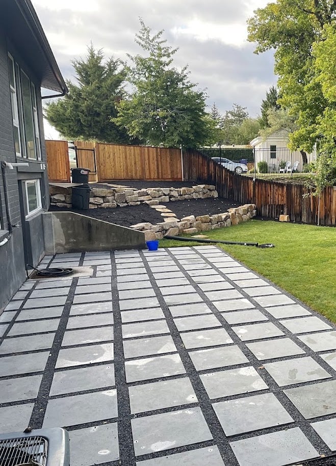Backyard with a grid-patterned stone patio, green lawn, terraced retaining wall with rocks, wooden fence, and trees in the background. Landscaping contractors in Holladay, Utah can help create a similar inviting outdoor space. A blue bucket is on the patio.