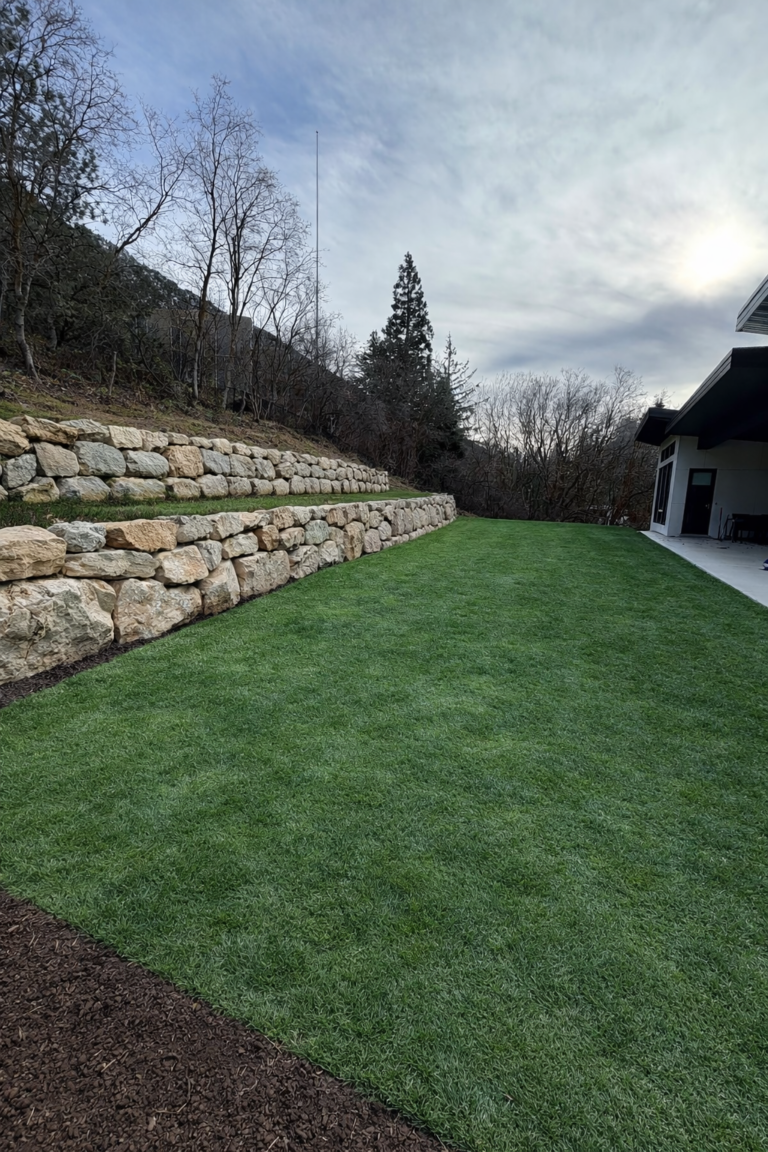 A neatly mowed lawn bordered by a stone retaining wall expertly crafted through skilled hardscape installation, with trees and bushes in the background and a house on the right.