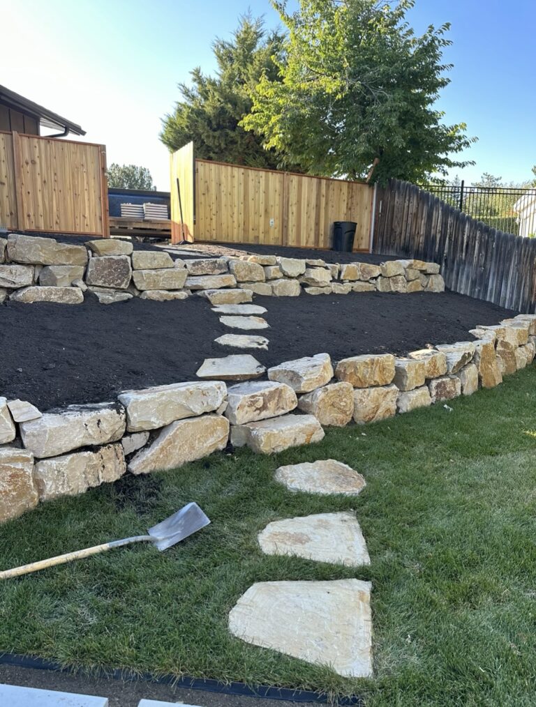 Stone steps lead up a terraced garden with large rock retaining walls, fresh soil, and a wooden fence—showcasing expert hardscape installation. A shovel lies on the grass near the steps, hinting at the thoughtful hardscape design behind this inviting outdoor space.