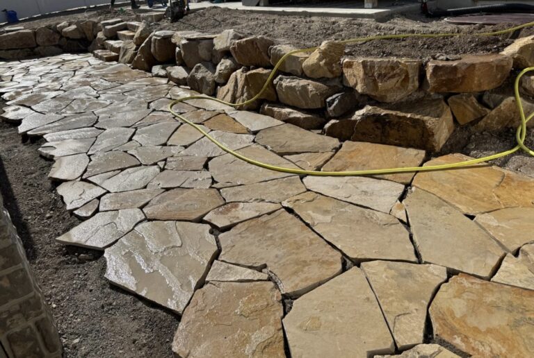 A beautifully crafted stone pathway and retaining wall, showcasing expert hardscape installation with large, irregularly shaped rocks, complemented by a yellow hose lying on the ground.