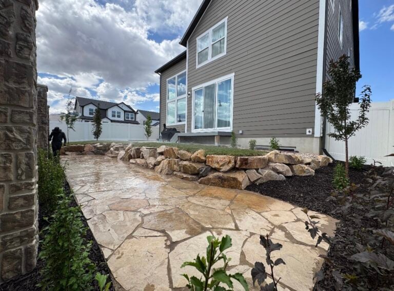 Stone patio with expert hardscape design and a retaining wall of large rocks beside a modern gray house; greenery lines the stone path, and a cloudy sky is visible overhead.
