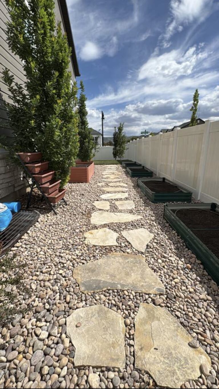 Stone pavers form a path through a gravel garden beside a house, showcasing expert hardscaping with raised garden beds and tall green shrubs along a white fence.