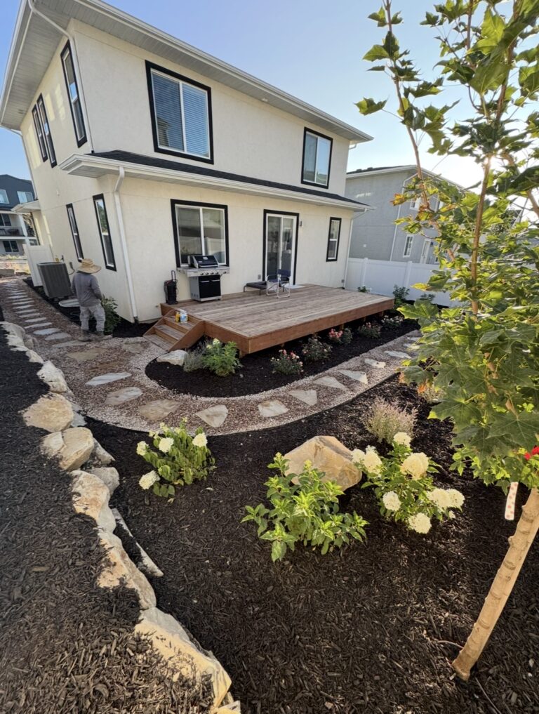 Two-story house with a wooden deck, stone path crafted through expert hardscape design, mulched garden beds, shrubs, and flowers in a landscaped backyard. A person stands near the side of the house.