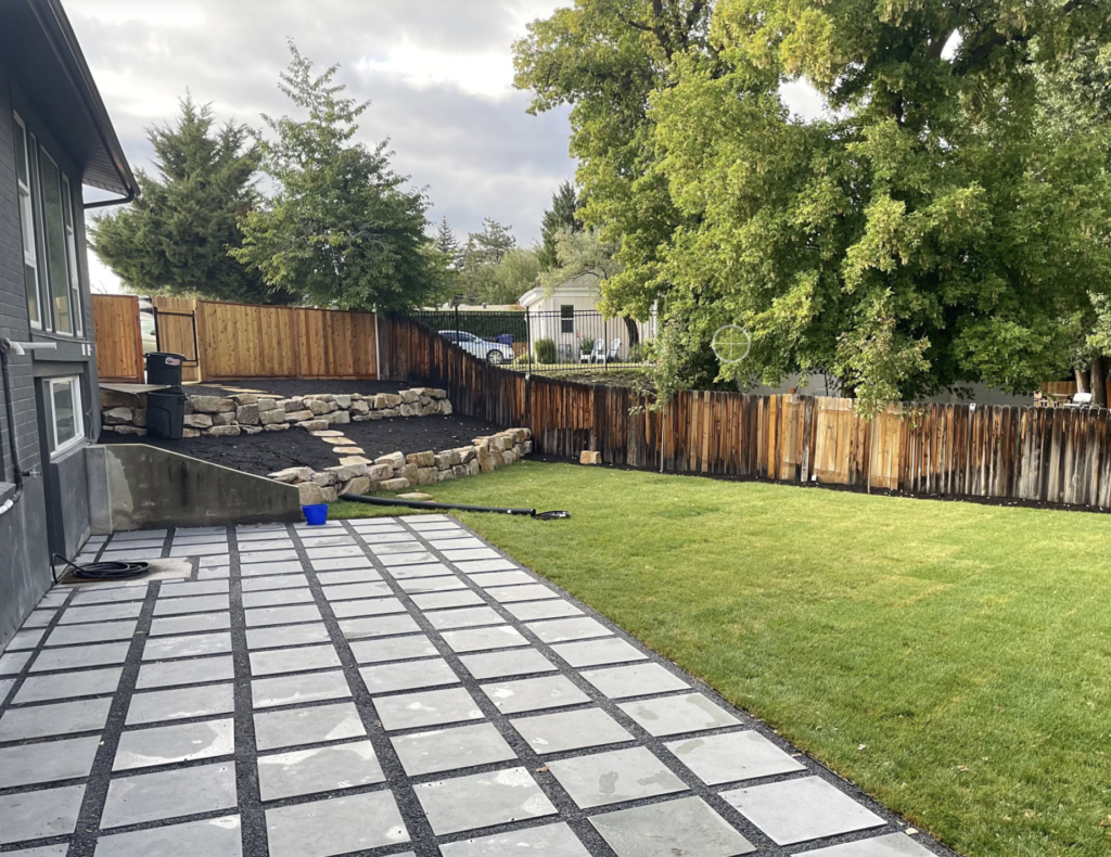 A backyard with a paved patio, green lawn, wooden fence, retaining wall with rocks, and mature trees along the fence line—perfect for showcasing our exceptional landscaping services.