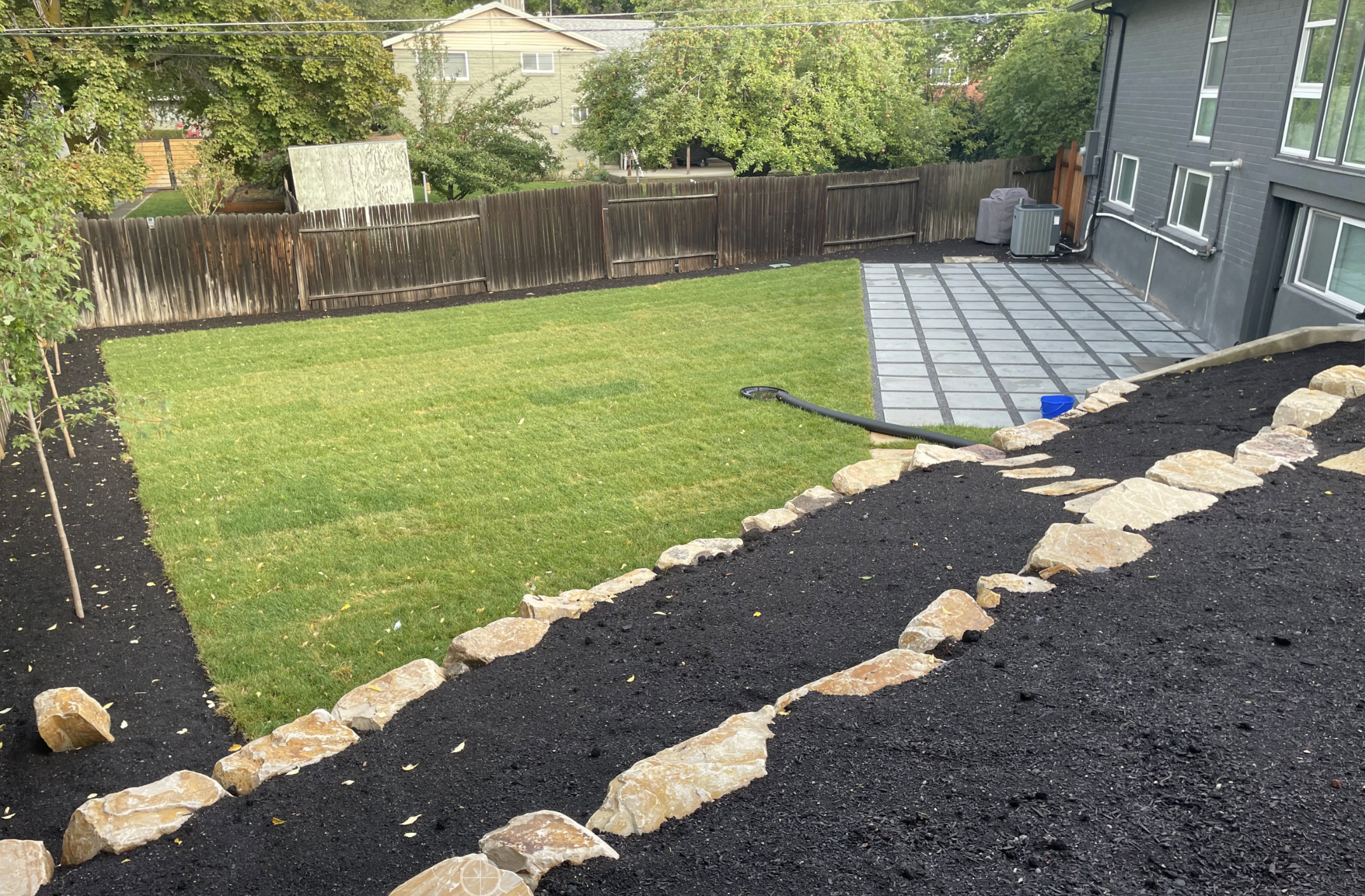 Backyard with a green lawn, paved patio area, rock-bordered pathway on a slope, and black soil surrounding the grassy area—see why our Landscaping Services are praised in Customer Reviews.