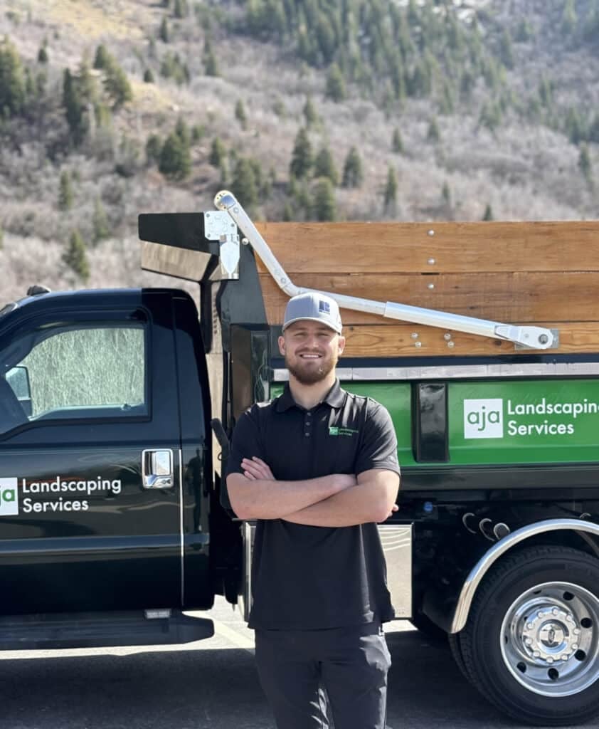 A person stands with arms crossed in front of a black truck labeled "aja Landscaping Services," set against trees and hills, perfect for the company’s Homepage.