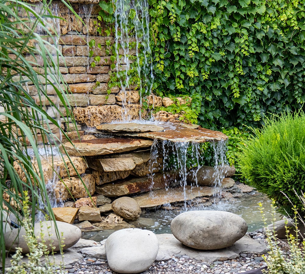 A small garden waterfall flows over stacked rocks, surrounded by green plants, an ivy-covered wall, and several large smooth stones—bringing a soothing homepage vibe to your outdoor space.