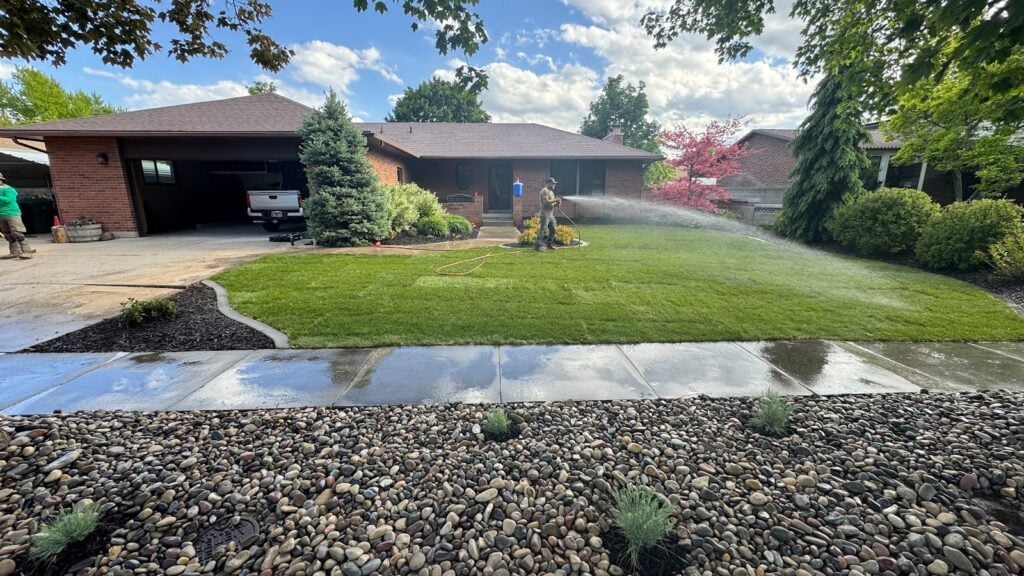 A sprinkler waters the front lawn of a brick house on a sunny day, showcasing beautiful landscaping. A person stands on the porch, and the driveway and sidewalk are wet. Shrubs and trees surround the yard, creating a lush, inviting atmosphere.