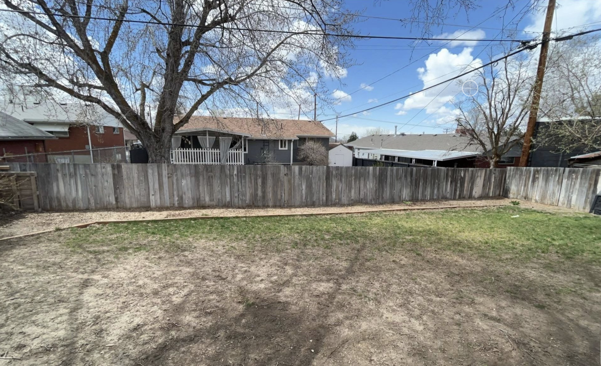 A backyard with a patchy lawn, bare soil, a wooden fence, and houses and trees in the background under a partly cloudy sky—perfect for showcasing landscaping services with impressive before-and-after transformations and customer reviews.