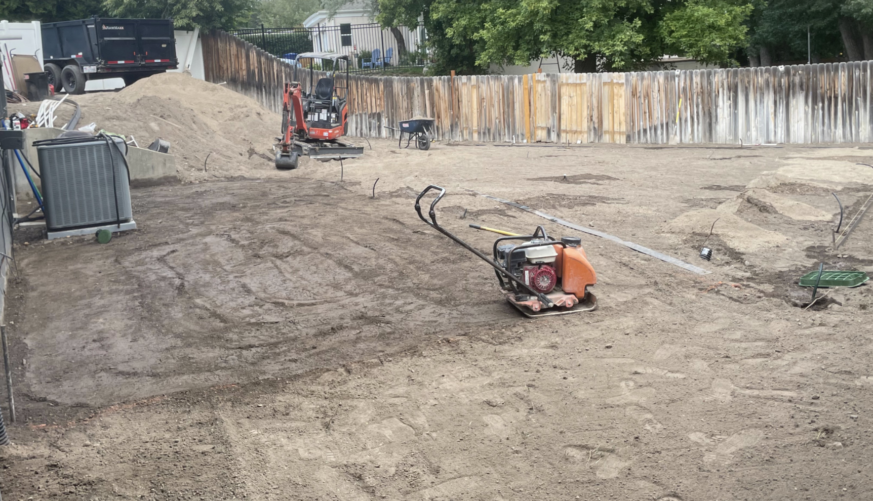 A backyard construction site with a soil compactor in the foreground, an excavator in the background, and piles of dirt along a wooden fence—perfectly showcasing the quality projects completed by AJA Landscaping Services.