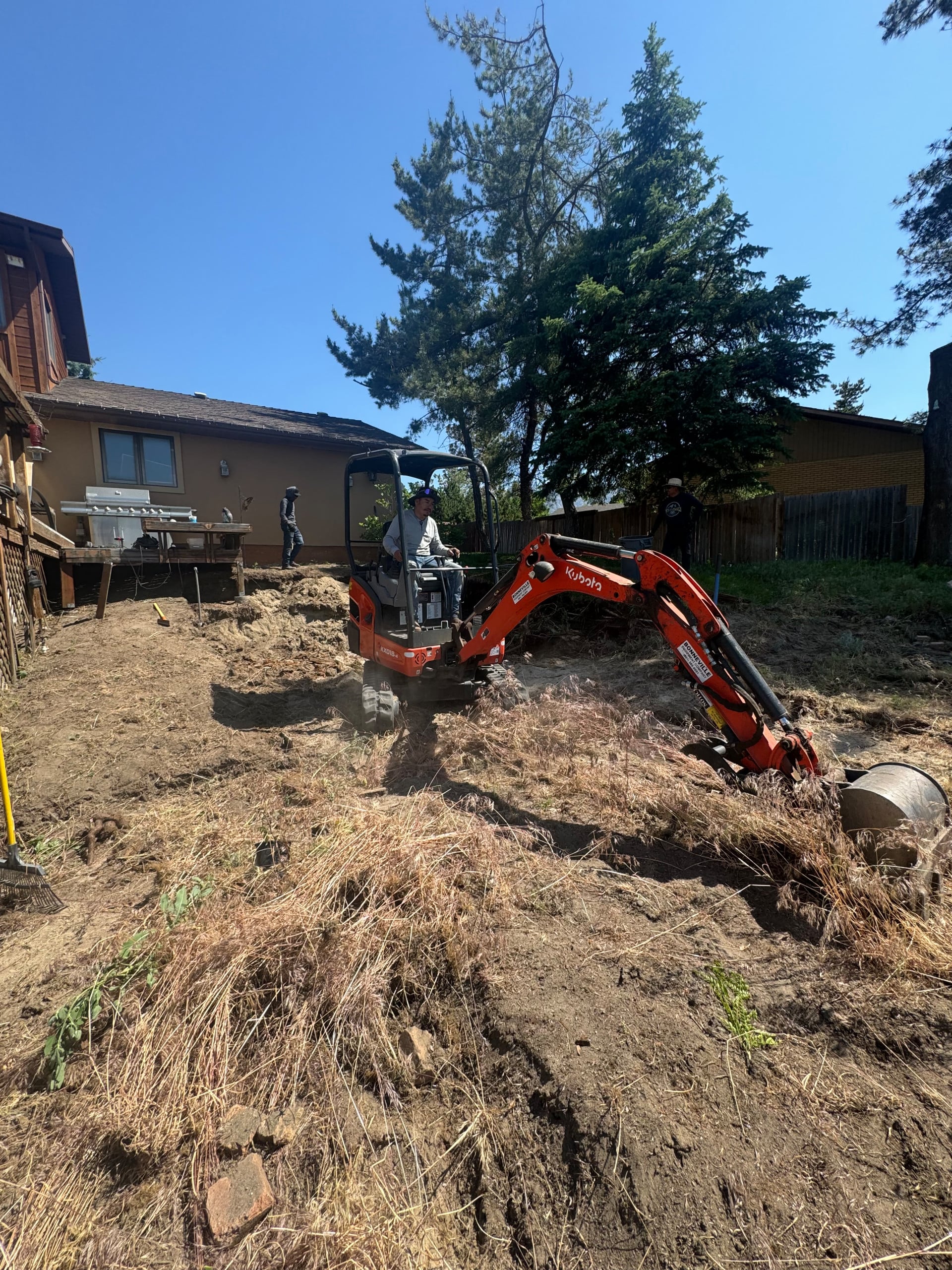 A person operates a small orange excavator on a dry, sloped yard beside a house, with another person standing nearby and trees in the background—a scene reflecting AJA Landscaping Services' dedication highlighted in customer reviews.