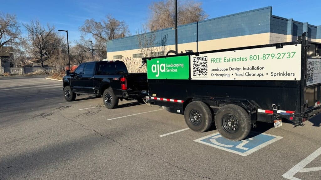 A black pickup truck with a landscaping trailer is parked in a handicapped space in a parking lot near a blue and beige building.
