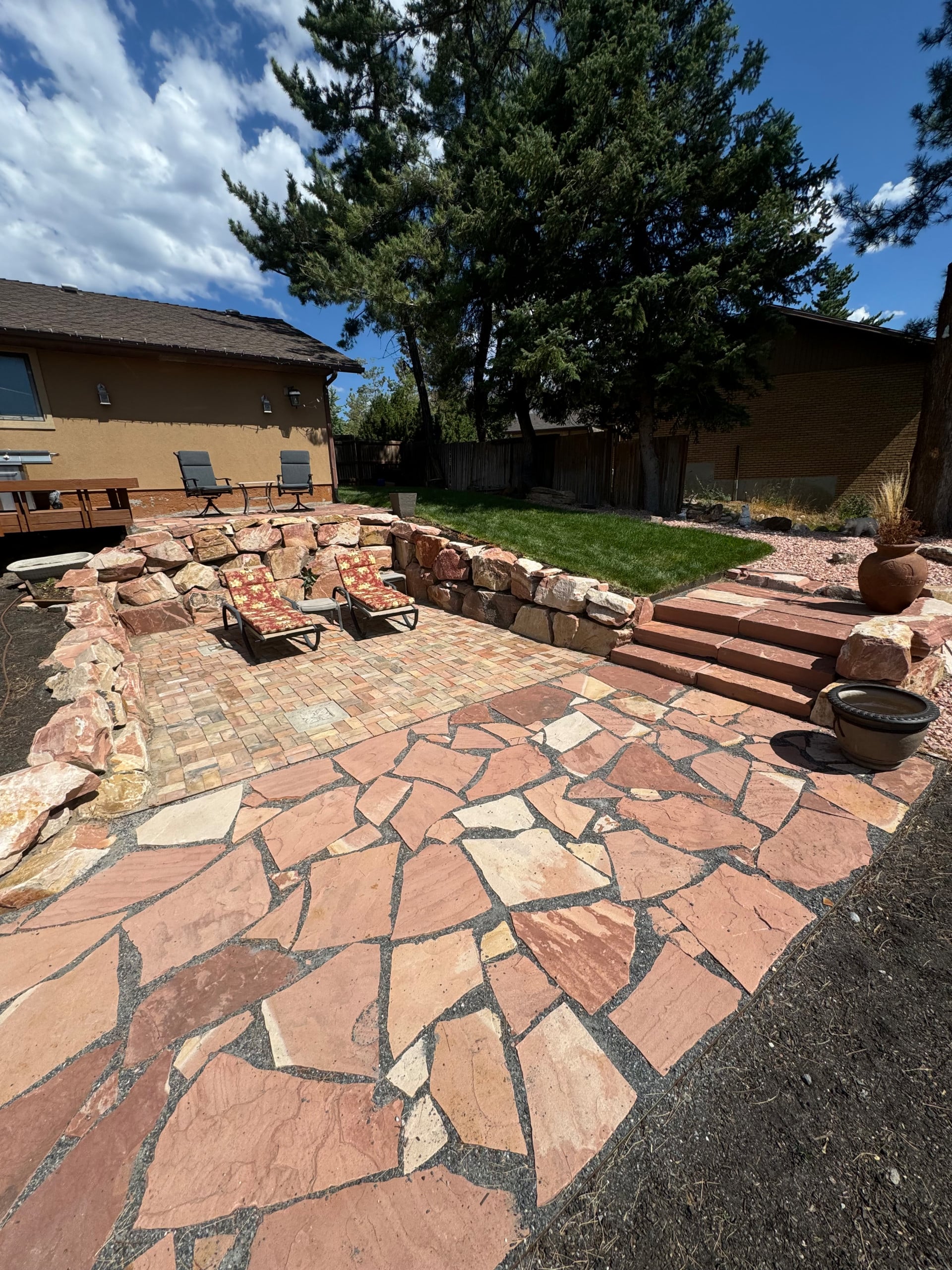 Backyard patio area with stone pavers, two lounge chairs, steps leading to a grassy yard, landscaping rocks, and trees in the background under a blue sky—discover why customers love it in our AJA Landscaping Services testimonials.
