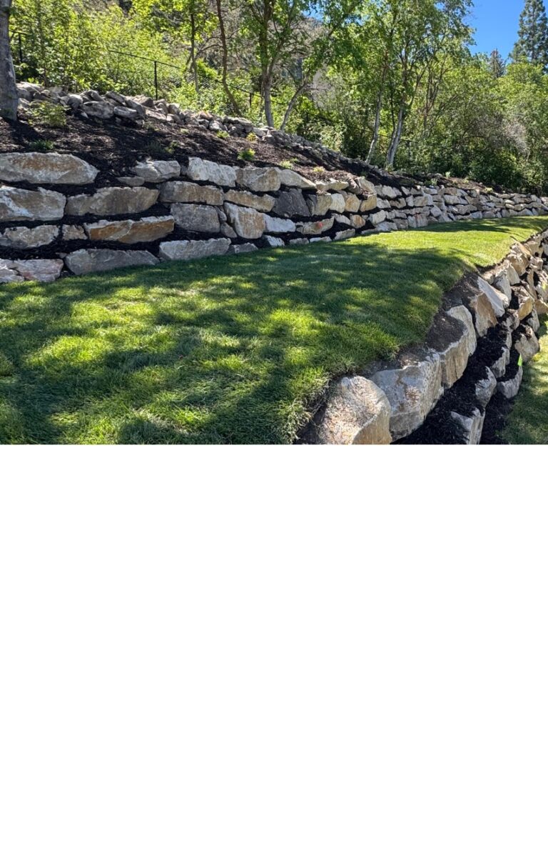Stone retaining walls with stacked rocks, crafted by a skilled hardscape company, border a neatly trimmed grassy area, with trees and shrubs in the background under sunlight.