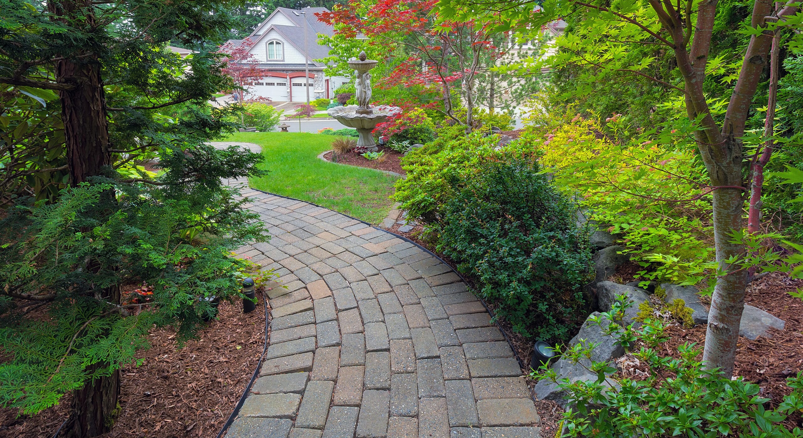 Curved stone pathway through a landscaped garden with green shrubs, trees, and a stone fountain in the background near residential houses, perfectly crafted by our expert Walkway Paver Installation service.